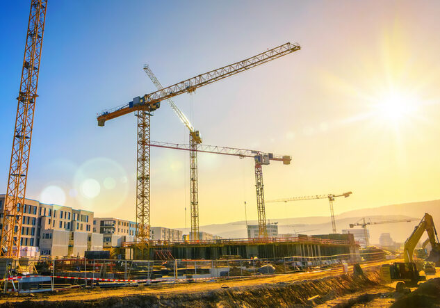 Large construction site including several cranes working on a building complex, with clear blue sky and the sun